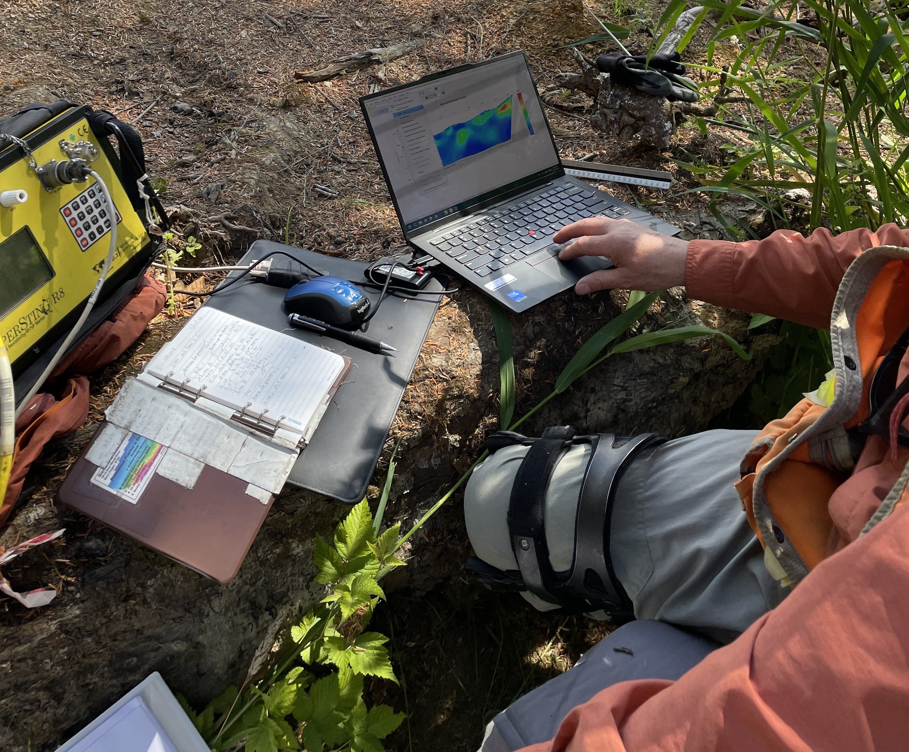 Field technician seated beside a creek bank in a wooded area, using a laptop to review a color-coded monitoring map while connected to specialized survey equipment.